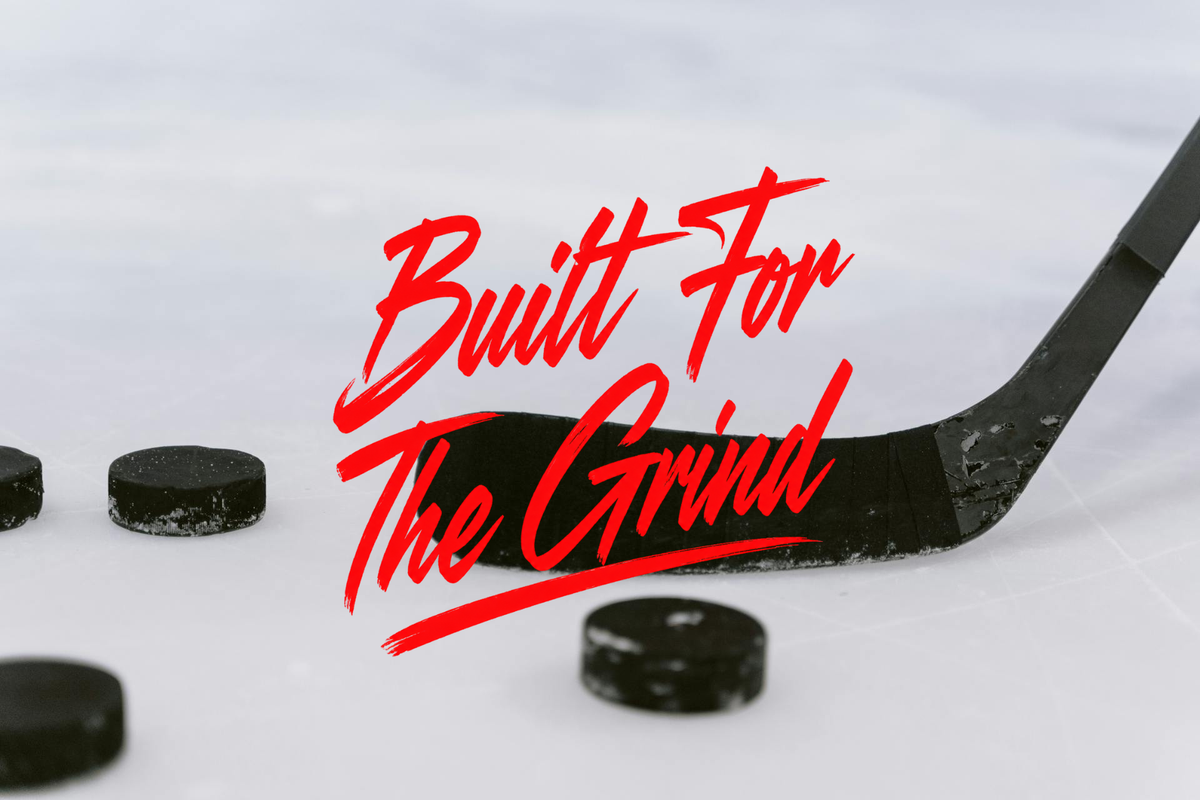A hockey stick and several black hockey pucks on an ice surface, overlaid with the red script text 'Built For The Grind'.